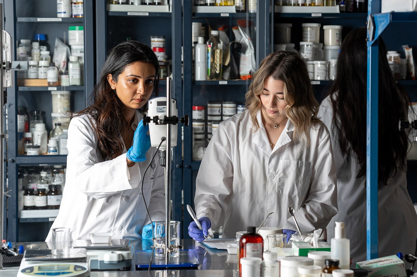 Two chemists in a cosmetic lab work with white coats and rubber gloves. Behind them, shelves display innumerable lab containers of creams, lotions and potions.