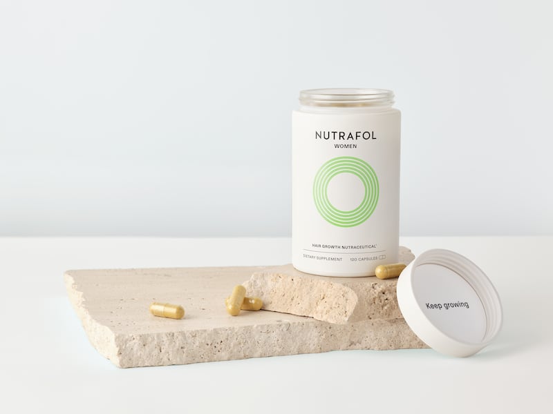 The image is a still life of the brand Nutrafol's hair growth supplements. The uncapped bottle sits on top of stacked stone tablets, which are littered with dull, golden capsules. The inside of the cap reads "Keep growing."