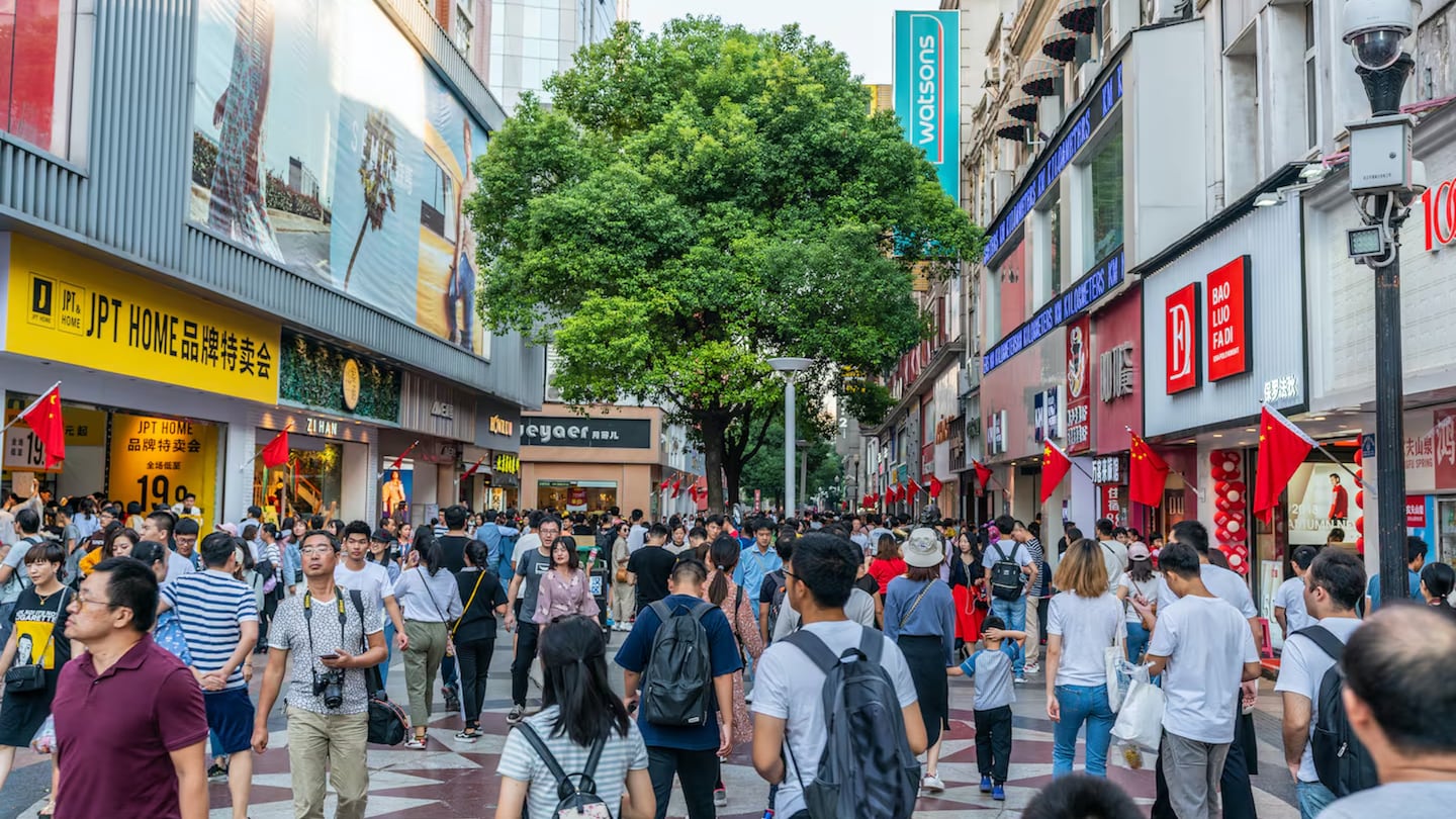 The image shows a lively urban street scene, likely in a shopping district. Numerous pedestrians are walking along the street, which is lined with modern storefronts featuring bright signage in both English and what appears to be Chinese characters. The architecture is contemporary, with large display windows showcasing various brands. Trees along the street add a touch of greenery, and the clear blue sky suggests pleasant weather. This scene captures the vibrancy and hustle of city life, highlighting the commercial and cultural activity in the area.
