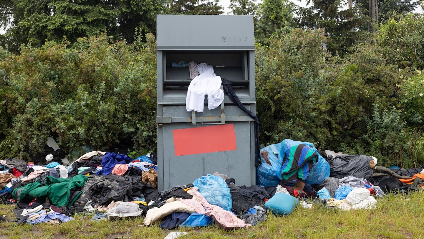A used clothing bin surrounded by piles of old clothes in a field.